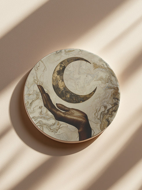 A ceramic coaster featuring a bronze-colored hand reaching toward an antiqued gold crescent moon against a swirling ivory and gold marble background, available in round and square shapes with cork backing.